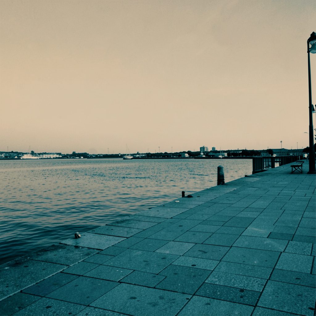 Hard two-tone duotone photograph of a calm harbor promenade with low skyline in muted warm tan and cool slate tones